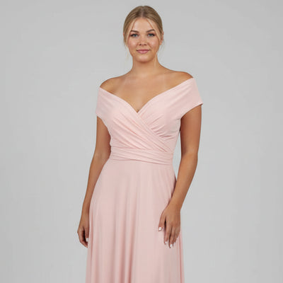 Woman wearing a pink off-shoulder dress standing indoors with decorative wall and plants in the background.